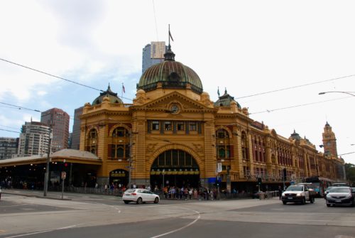 a large building with a dome and people walking in front of it with Flinders Street railway station in the background