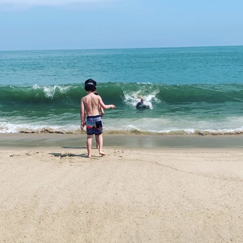 a boy standing on a beach looking at a person swimming in the water